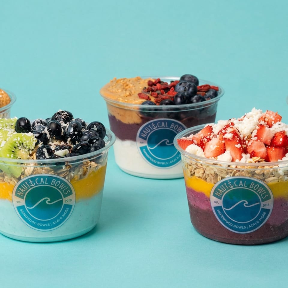 Five colorful Nautical Bowls smoothie bowls topped with fruit, granola, coconut, and nuts against a turquoise background.