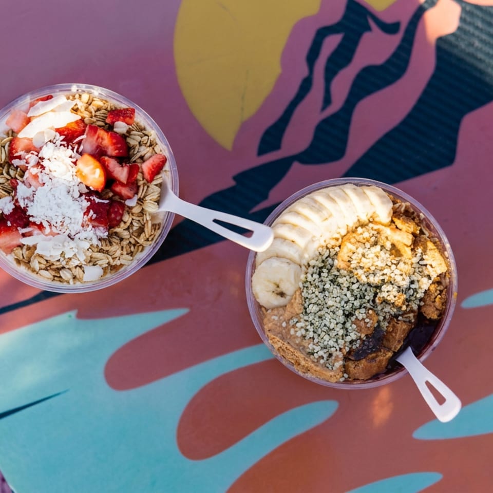 Two smoothie bowls topped with strawberries, banana, granola, coconut, and seeds sit on a colorful patterned table.