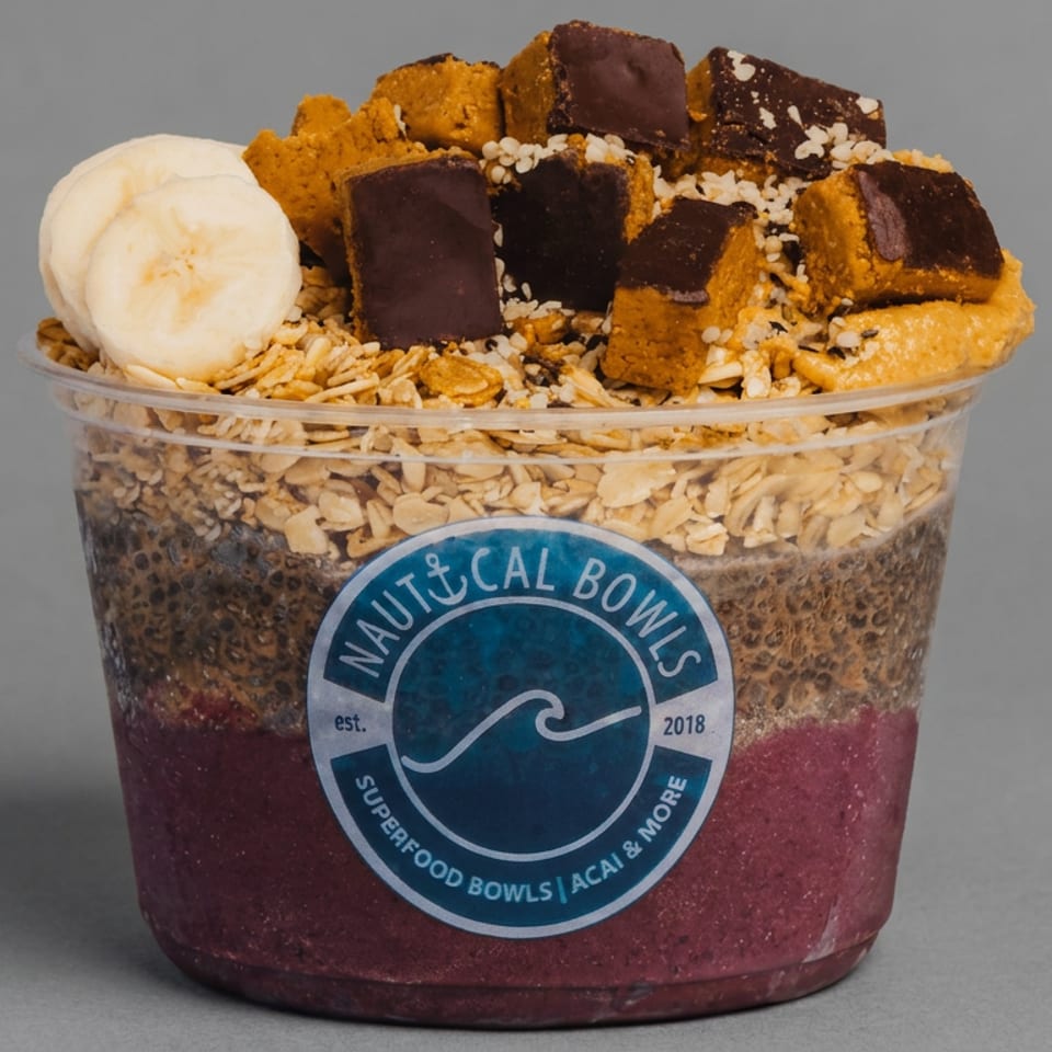 A layered Nautical Bowls acai cup topped with banana slices, granola, chocolate-covered pieces, and seeds against a gray background.